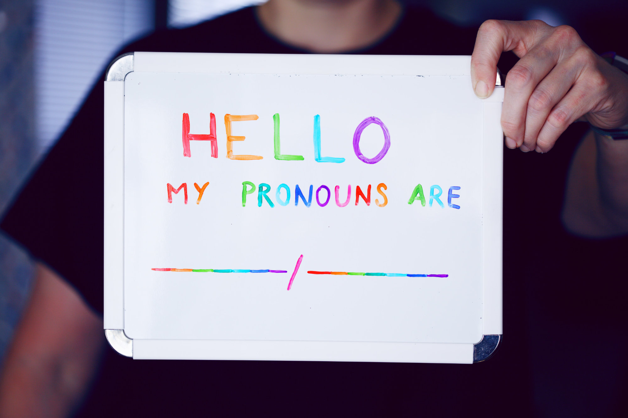 A person holds a whiteboard sign that reads, "Hello my pronouns are..."