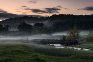 An early morning landscape, shrouded in fog, a bridge spans a small stream.