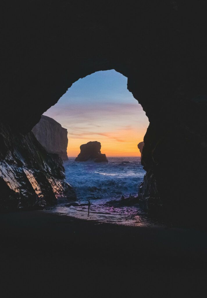 A sunset appears at the exit of a cave near the ocean.